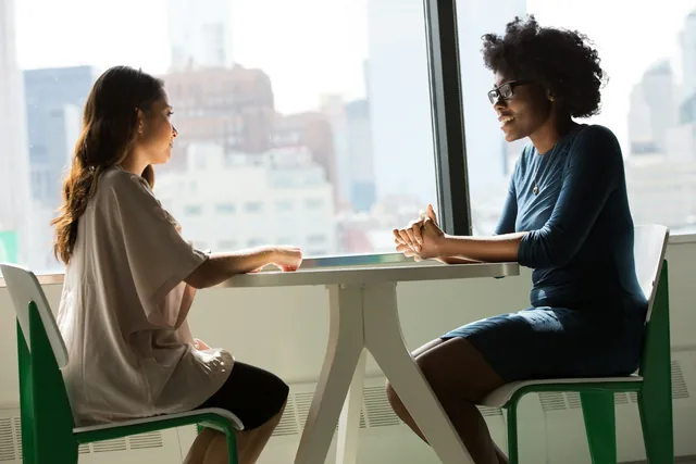 Two people talking across a table by a window